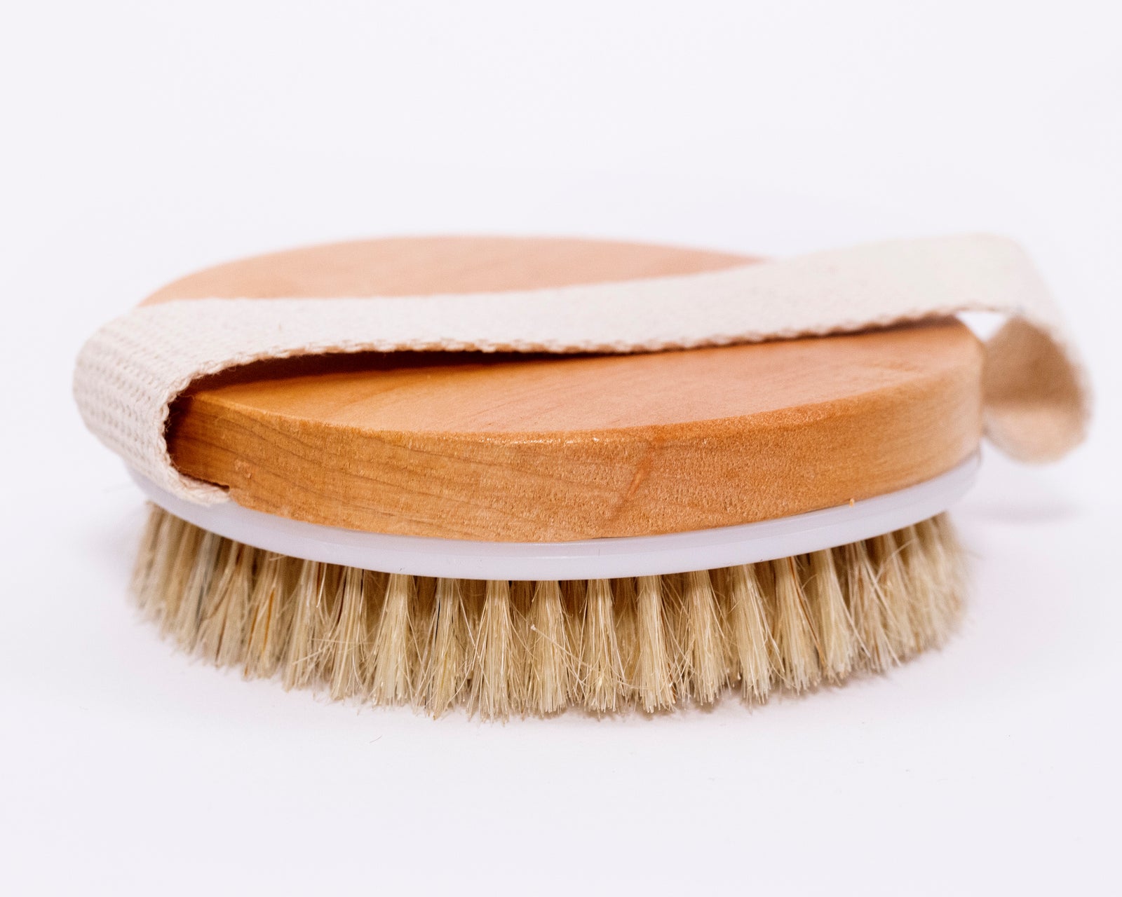 A round wooden brush with natural bristles rests on a white surface; a light-colored fabric strap encircles its base.