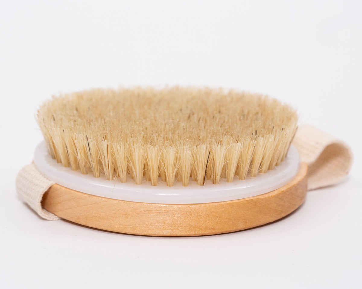 A round dry brush with natural bristles rests on a white background.  It has a wooden base and a light-colored fabric strap. The brush is positioned to show its side profile.
