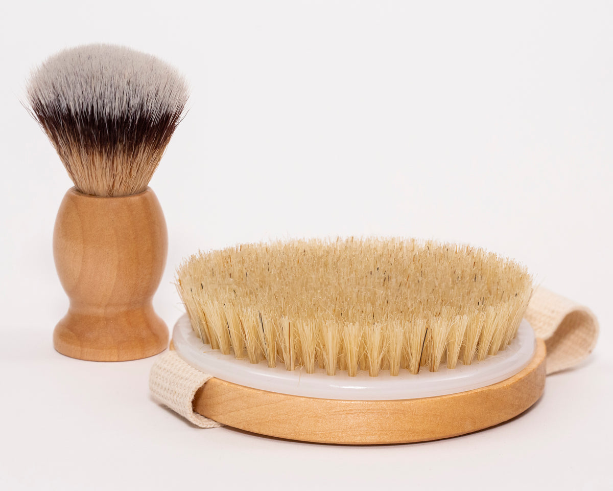 A shaving brush and a body brush rest on a white surface.  The shaving brush has a wooden handle and badger hair bristles; the body brush has a wooden handle, a white center, and natural bristles with a fabric loop.